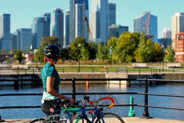 femme debout, la main sur la selle de son vélo accoté contemple la ville de Montréal. Les arbres sont verts et les bâtiments reflètent au soleil